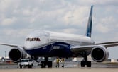 Two men wearing safety vests walk near a Boeing 787-10 Dreamliner on a runway.