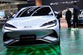 A sleek, light-silver car displayed on a green patterned showfloor. A woman and two men inspect the car. The license plate holder says "BYD Sealion." Behind the car are two covered vehicles with the words "BYD | REVER Automotive" on the wall.