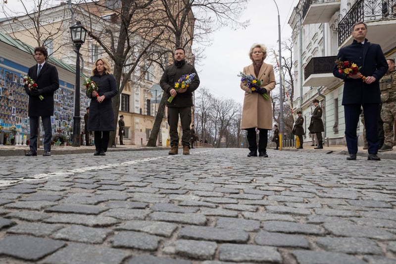 Five world leaders walk with bouquets on a cobbled street.