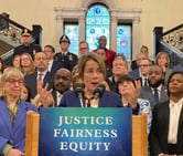 Maura Healey speaks during a news conference, with more than two dozen people standing on a staircase behind her.