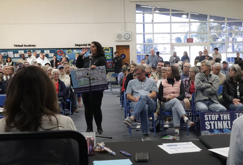 A woman, holding a map with the text "keep the sewage in Mexico," speaks in front of a microphone at a well attended public meeting.