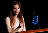 Amanda Knox points with her right hand while giving a speech from behind a lectern.