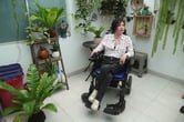 A woman sits in a wheelchair in a room filled with plants.