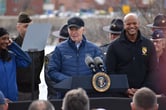 An elderly man in a black hat and a windbreaker stands in front of a podium.