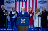 Joe Biden gestures with his hands while speaking during a campaign event, with several members of the Kennedy family applauding behind him.