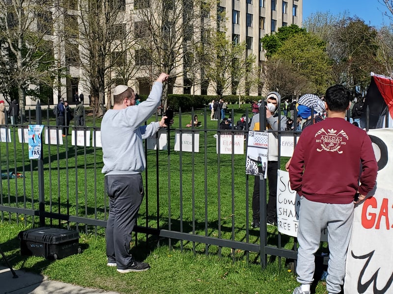 A man stands in front of a fence and points at another man on the other side