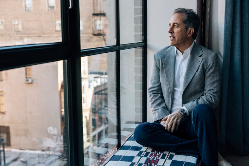 Jerry Seinfeld looks out a window while sitting down.
