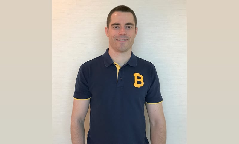 A headshot of Roger Ver standing against a beige wall wearing a navy blue polo T-shirt with a yellow B on the left breast.