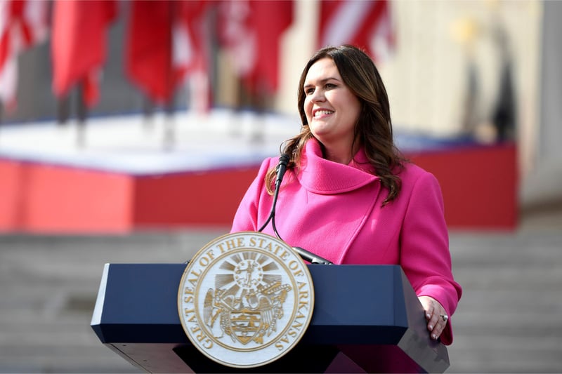 Sarah Huckabee Sanders smiles while standing behind a podium outdoors.