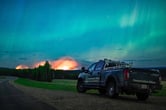 A pick-up truck parked in a field, with flames and smoke from a fire rising in the distance and Aurora Borealis in the sky.