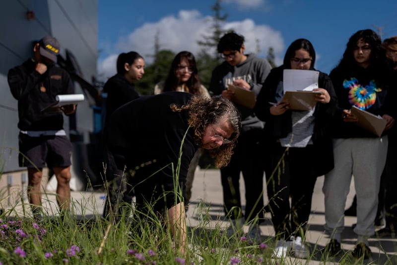 A teacher leans over to examine plants as students holding clip boards review their notes.