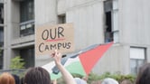 A person holds a beige sign that reads "our campus" black letters with a red underline under the word "our." A black, red, white, and green Palestinian flag can be seen in the background.