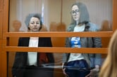 Zhenya Berkovich and Svetlana Petriychuk stand inside a mostly glass cage in a courtroom.