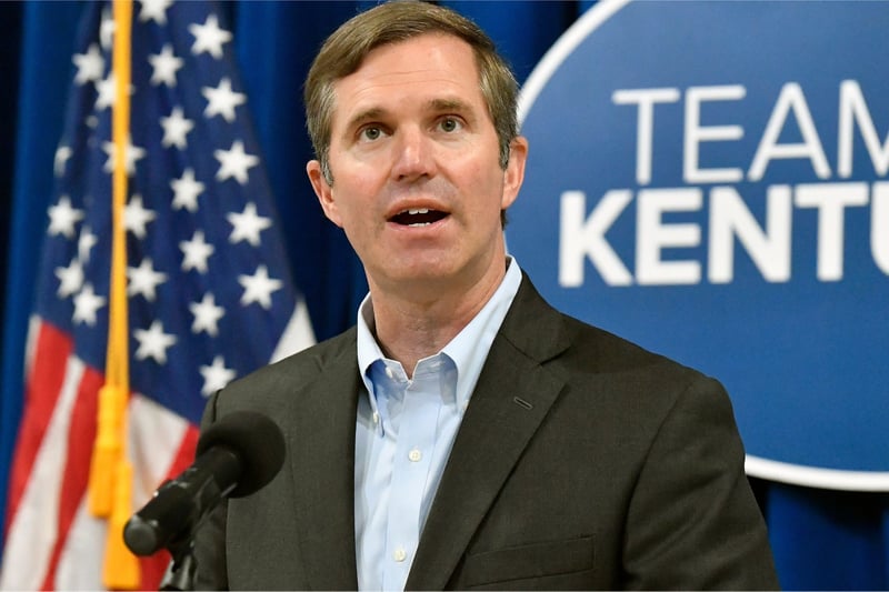 Wearing a dark gray blazer over a light blue button-down shirt, Andy Beshear speaks into a microphone while standing in front of an American flag and a partially visible sign with the letter "team" above the letters "kent."