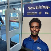 A man wearing a striped Under Armour collared shirt and khaki pants walks out of a Best Buy store near a hiring sign showing a young man wearing a Best Buy polo shirt, with the text "now hiring" and "Apply in store or at jobs.bestbuy.com" next to a QR code and the Best Buy logo also on the sign.