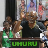 An older Black man gestures with a pencil in his right hand while standing behind a podium with two microphones pointed at it and "Uhuru" in green letters on the front, with nine people and separate images of three people and a white sedan in a field behind him.