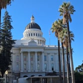 A large building with four distinct columns stands tall against a blue sky, with palm trees lining its path.