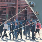 A group of people walk, being led by security guards. The front of a docked boat can be seen in the foreground.