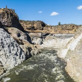 Steep canyon-like walls, carved out or blasted long ago, surrounds swiftly moving water, with blue sky and white clouds above.