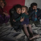 Two young Palestinian children cuddle near two other children as they sit on dirt with debris in it.
