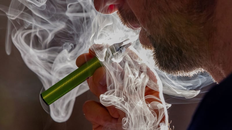 A man's face from nose to chin, showing a light beard, is surrounded by smoke curls from a green vape pen held near his mouth.