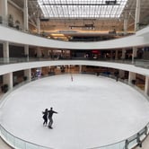 A couple in ice skates clasp hands in a large oval ice inside of a mall.