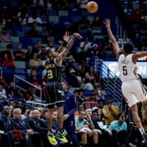A player wearing a dark Charlotte Hornets jersey shoots over a player wearing a mostly white New Orleans Pelicans jersey during an NBA game, with coaches, other players and part of the crowd in the background.