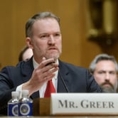 A man gestures with his right hand while holding a pen, with two water bottles, a name placard with, "Mr. Greer" in black text and a microphone on the table in front of him, with three people sitting behind him.