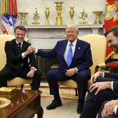 Emmanuel Macron laughs and Donald Trump smiles as they bump fists while seated in gold armchairs, with gold ornaments on a mantle behind them. JD Vance and Marco Rubio smile on one side as a woman in black looks toward them.