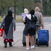 A woman carrying a child in winter clothes walks behind a person pulling two suitcases and a woman in a long head covering down a concrete road.