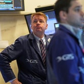 A man in a suit and striped tie puffs up his cheeks and purses his lip, hand on hip. Another man walks past him and multiple monitors and pieces of paper displayed on a wall.