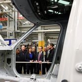 Keir Starmer stands near the doorway of a rough automobile frame inside a factory with a group of other people.