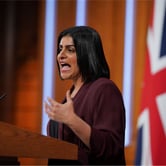 A woman wearing a maroon blazer over a black top gestures with her left hand while speaking from behind a podium.