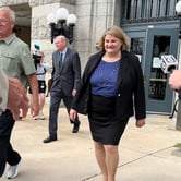 A woman in a skirt suit walking out of a building surrounded by men in suits while news cameras and microphones point toward her.