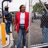 LaMonica McIver, in a red blazer, white shirt and jeans, walks through a tall chain link fence with two guards nearby.