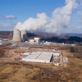 Steam rises from two cooling towers at a nuclear power plant next to hills, buildings and dozens of parked vehicles.
