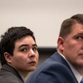 A young man in a gray suit sits beside a man in a blue suit inside a courtroom, both looking over their shoulders