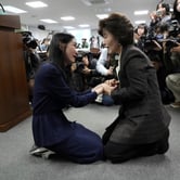 Two women kneel on the ground in a room full of people and overhead lights, holding hands, as photographers take their picture.