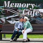 An elderly couple kisses while sitting on a bench in front of a large outdoor sign reading “Memory Café.”