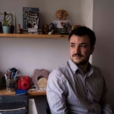Mahmoud Khalil sits at a desk with items on two shelves behind him.