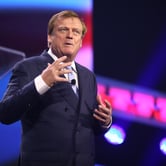 Man in a dark suit speaking on stage with colorful lights in the background.