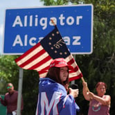 A woman waves an American flag in front of a blue and white road sign reading "Alligator Alcatraz" while another woman in a Make America Great Again cap stands nearby, draped in a red, white and blue cloth.