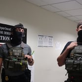 Plainclothes federal agents in gray and black plainclothes, wearing black face masks and various federal law enforcement vests, stand in the hallway outside of an immigration courtroom in New York City, while press photographers take photos of the arrest.