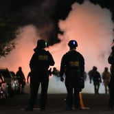 Three law enforcement officers stand on a street at night as smoke fills the background during a protest.