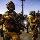 Troops in camouflage with facemasks and guns stand in the street at dusk.