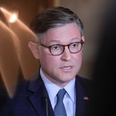 A man wearing glasses and a navy blue suit speaks at a podium, appearing serious as he addresses an audience.