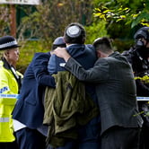 Men in yarmulkes hold one another around the shoulders behind a crime scene tape as police work outside.