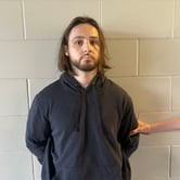 A young man with a beard, mustache and long brown hair stands against a white cement-block wall in a dark hoodie while someone holds his arm from outside the frame.