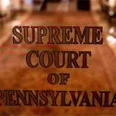 A glass door with the words “Supreme Court of Pennsylvania” etched on it, with a carpeted hallway and warm lighting visible in the background.