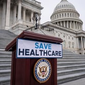 A podium with a “Save Healthcare” sign stands on the steps of the U.S. Capitol, with the building’s dome and columns visible in the background.